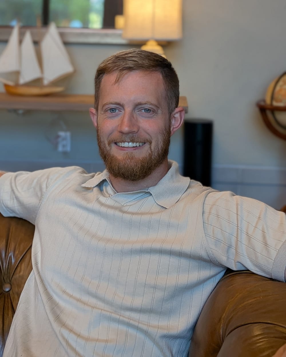 Jonathan Engle relaxing on a leather sofa, smiling, with a model sailboat on the shelf behind him